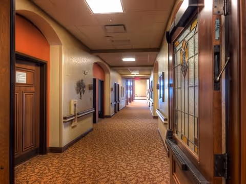 A well-lit hallway in a senior living facility with patterned carpet, beige walls, handrails on both sides, and several closed wooden doors. The hallway extends into the distance with ceiling lights and decorative wall art.
