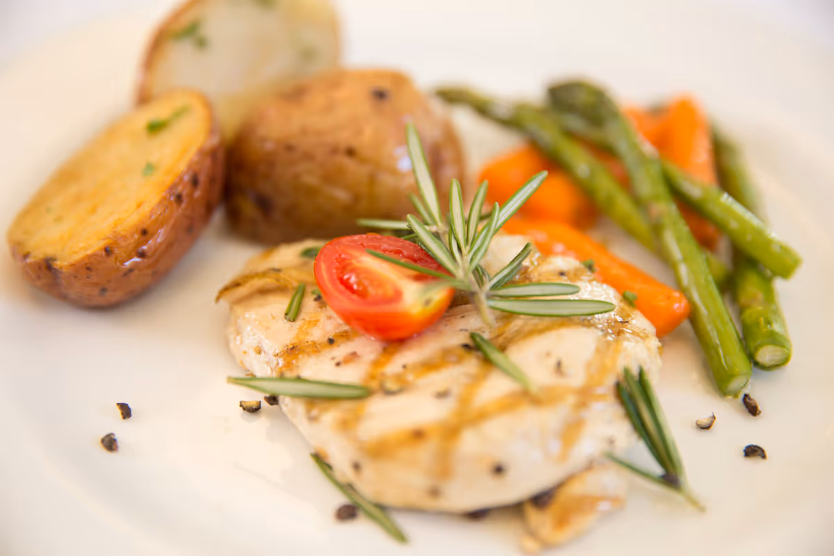 Grilled chicken breast garnished with rosemary and a cherry tomato, served with roasted potatoes, asparagus, and carrots on a white plate.