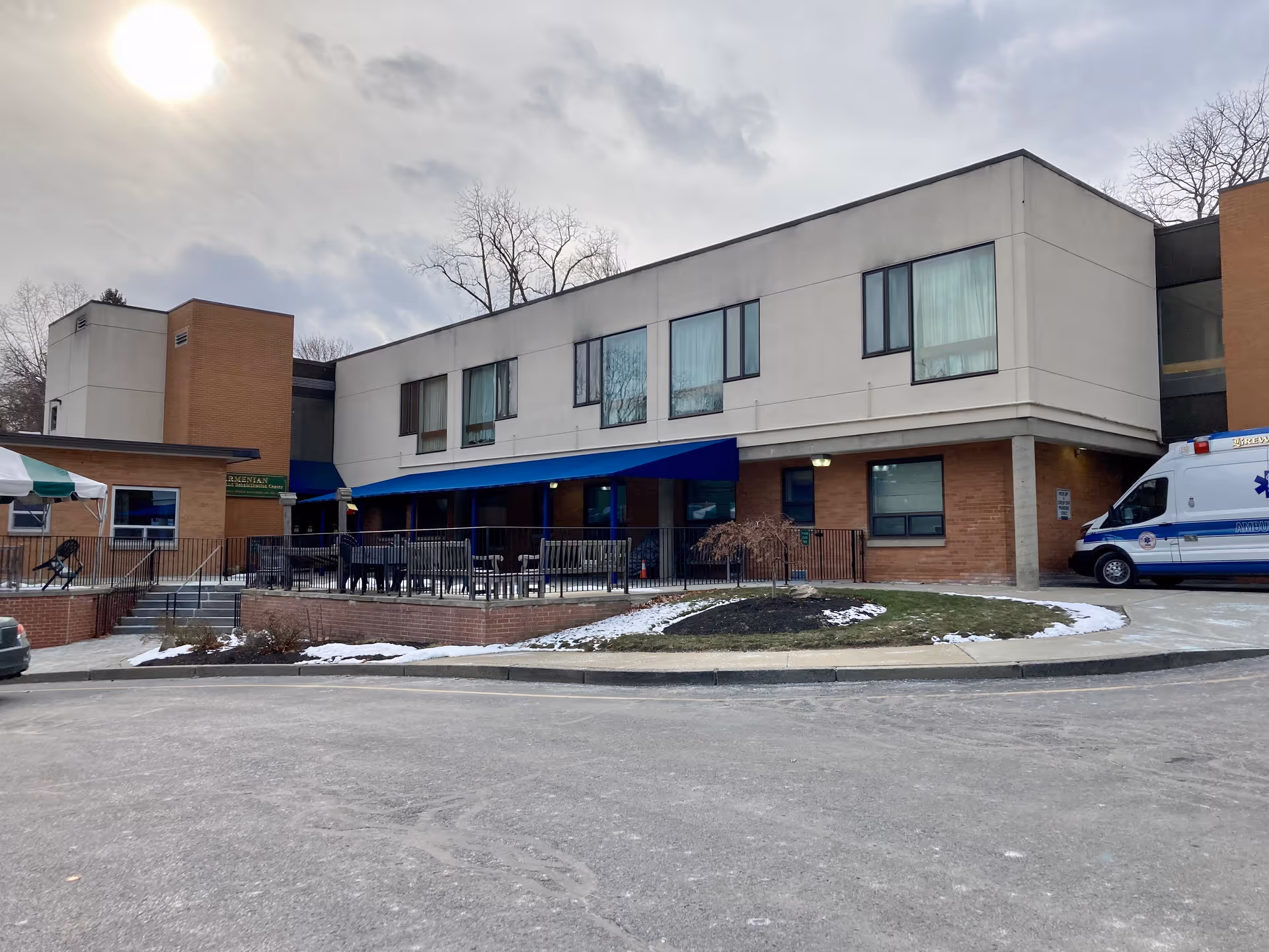 Exterior view of a two-story nursing and rehabilitation center building with a brick and white facade. There is a blue awning over a patio area with tables and chairs. An ambulance is parked on the right side of the building. The sky is cloudy with the sun partially visible.