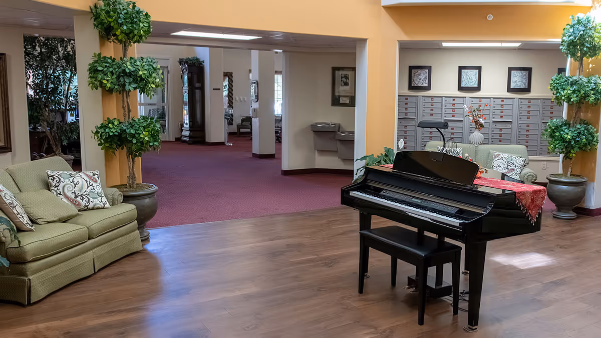 Communal lounge area with a black grand piano, seating, and potted plants in a senior living facility.