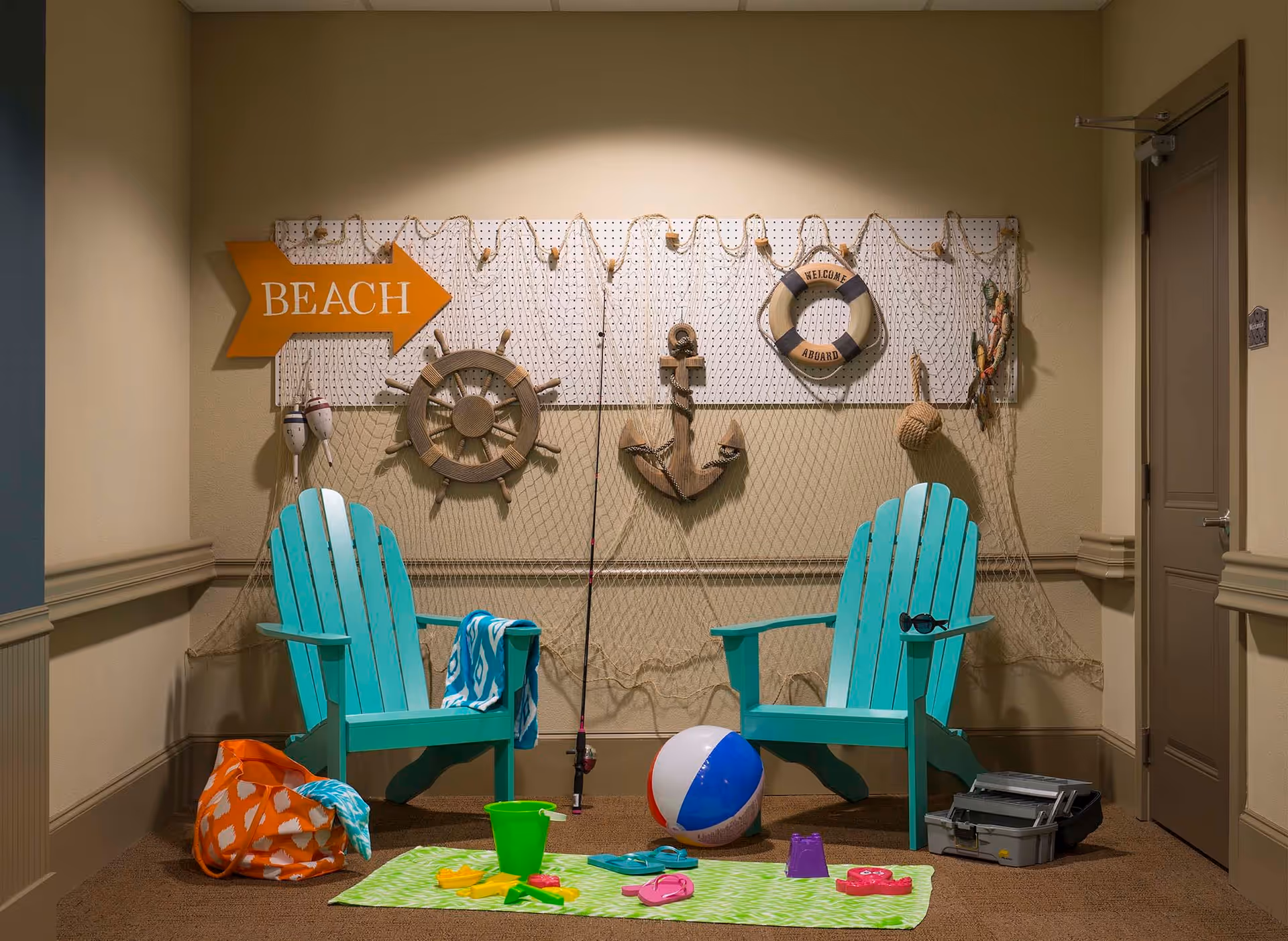 Indoor beach-themed seating area with two turquoise Adirondack chairs, a fishing pole, a beach ball, a green bucket, sand toys on a green towel, and a decorative wall with a fishing net, wooden ship wheel, anchor, life preserver, and a sign pointing to the beach.