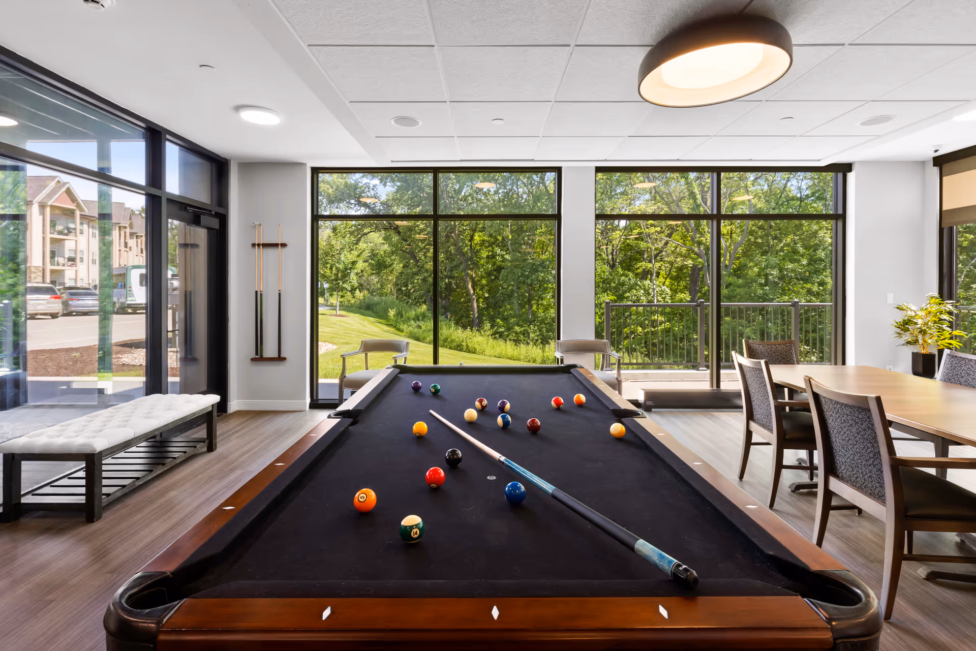 A bright and modern common area with a black felt pool table in the center, pool cues mounted on the wall, a bench near the glass entrance door, and a dining table with chairs near large windows overlooking green trees outside.