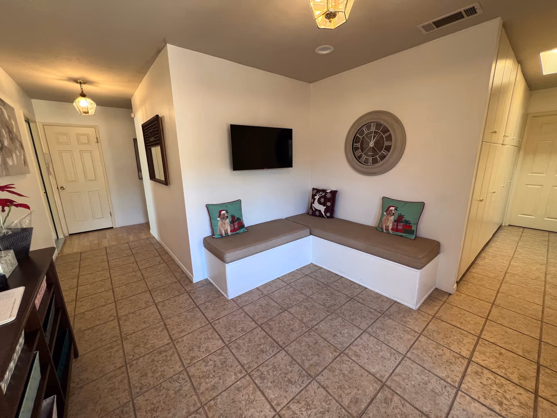 Small entry lounge with an L-shaped built-in bench topped with cushions, a wall-mounted TV and a large clock on a tiled floor.
