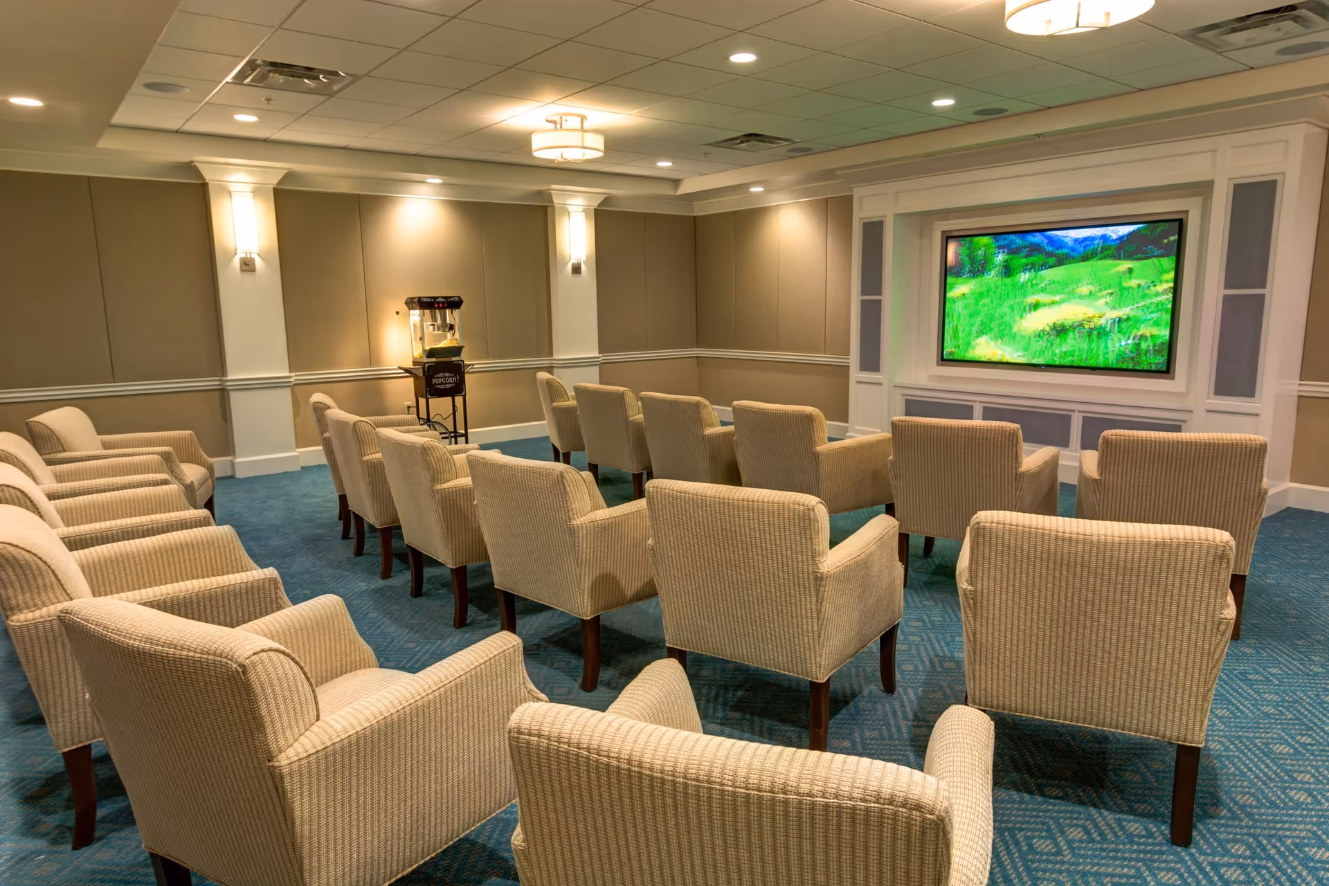 A cozy media room with multiple beige upholstered armchairs arranged in rows facing a large flat-screen TV mounted on a white paneled wall. The room has soft lighting, blue carpet, and a popcorn machine in the corner.