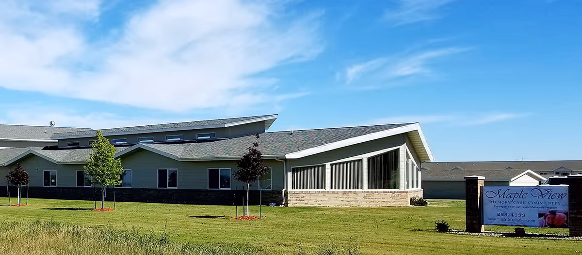 Exterior view of a single-story building with a sloped roof, several windows, and a well-maintained lawn with small trees. A sign in front reads Maple View Memory Care Community with contact information.