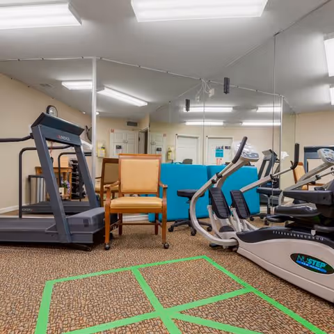 Interior view of a fitness room in a senior living facility with exercise equipment including a treadmill and a NuStep recumbent cross trainer. There are two chairs, one orange and one blue, positioned near the equipment. The room has a carpeted floor with green tape markings and a large mirror covering one wall reflecting the equipment and chairs.
