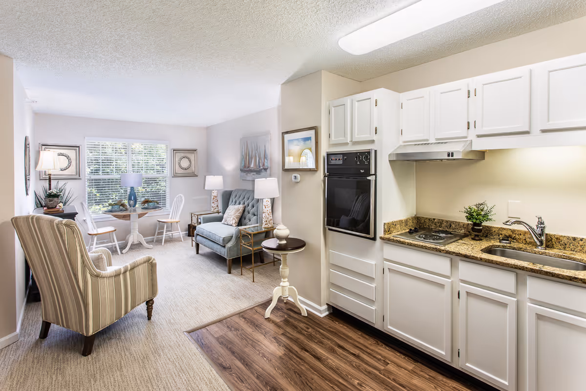 Bright open-plan interior showing a small kitchen with white cabinets and a cozy living area with chairs and a dining table by a window.