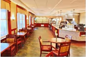 A spacious dining area with multiple wooden tables and chairs arranged neatly. The room has large windows on the left side allowing natural light to fill the space. On the right side, there is a long counter with food service equipment and pendant lights hanging from the ceiling. The floor has a patterned design, and the walls are painted in warm tones with decorative wall sconces.
