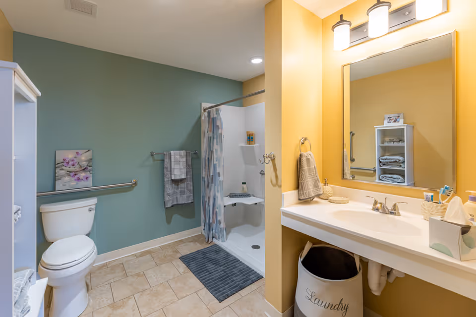 A clean and accessible bathroom with a white toilet, a walk-in shower with a curtain, and a large mirror above a white sink countertop. The walls are painted green and yellow, and there are towels hanging on metal bars. A white shelving unit holds folded towels and a small picture frame. A laundry basket is placed under the sink.