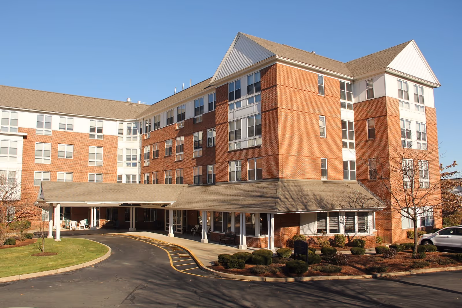 Brick multi-story senior living building with a covered driveway entrance and landscaped grounds under a clear blue sky.