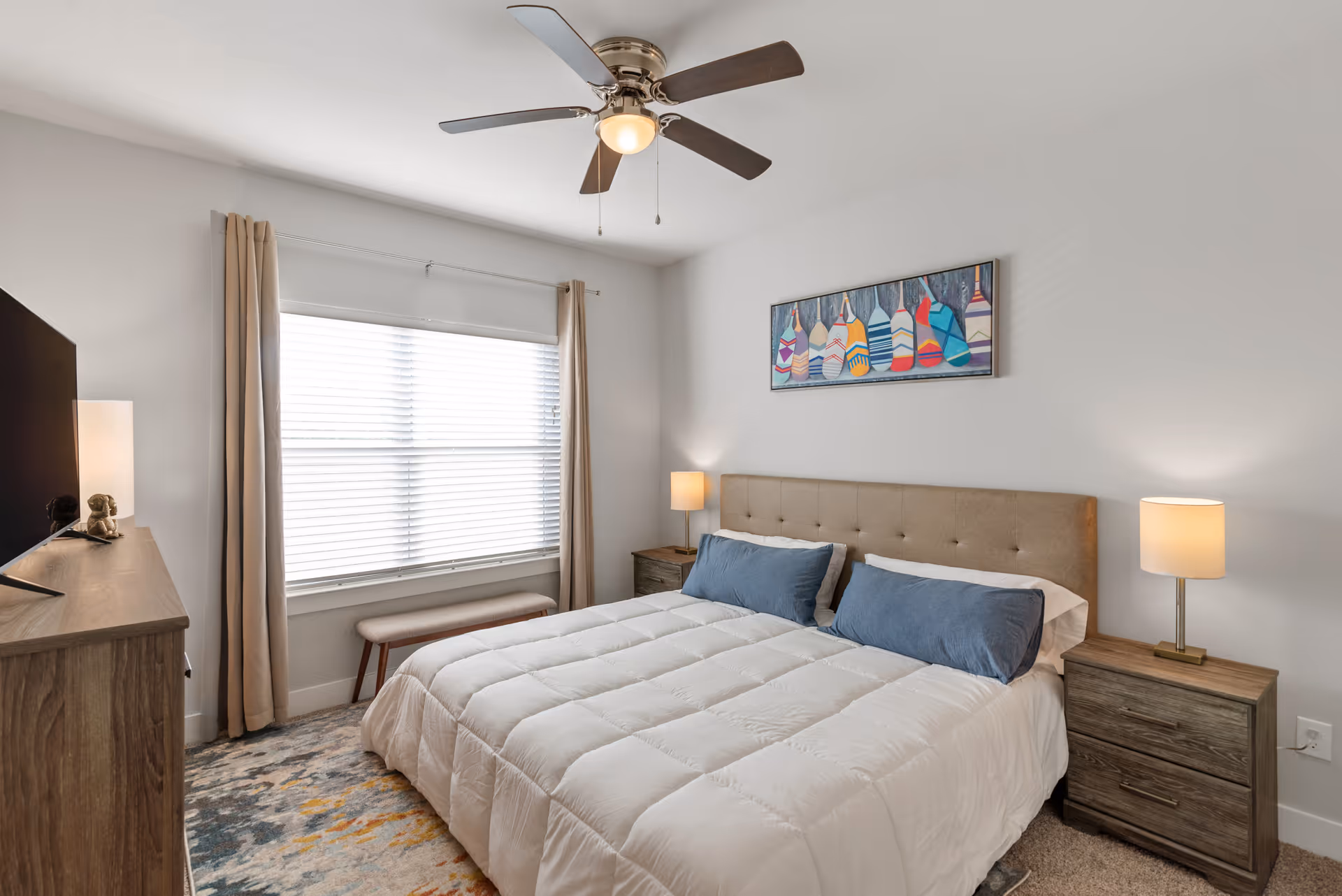 Bright, neatly styled bedroom with a bed, upholstered headboard, bedside tables and lamps, ceiling fan, window with curtains, dresser and wall art.