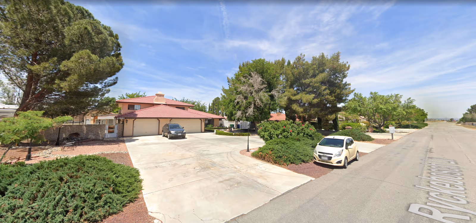 Exterior view of a residential building with a red-tiled roof and a three-car garage. There are two cars parked in the driveway and on the street. The property is surrounded by trees, bushes, and landscaped areas with gravel. The sky is clear with some wispy clouds.