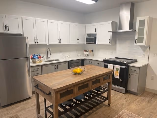 Modern kitchen with white upper cabinets and gray lower cabinets, stainless steel refrigerator and stove, a wooden island with drawers and a bowl of lemons on top, and a white tile backsplash.