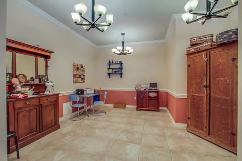A room with beige tiled flooring and walls painted beige with a red lower half. The room contains wooden furniture including a large cabinet on the right, a sideboard with a mirror on the left, and a smaller cabinet against the far wall. There is a small table with two chairs near the left side and a wall-mounted shelf with decorative items. Two ceiling light fixtures with multiple bulbs hang from the ceiling.