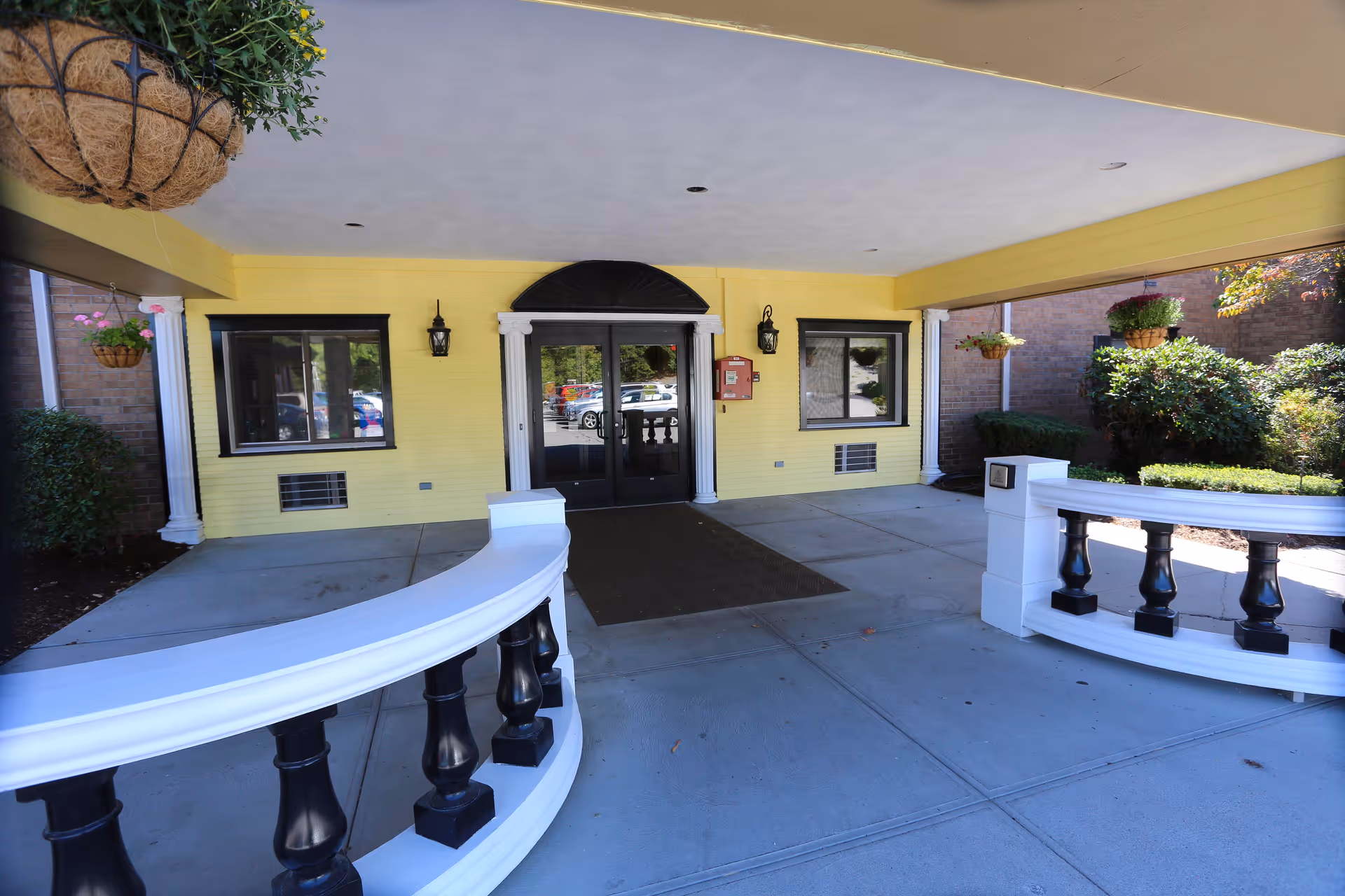 Covered main entrance with double glass doors set in yellow siding, flanked by windows, hanging planters, and curved white balustrades.