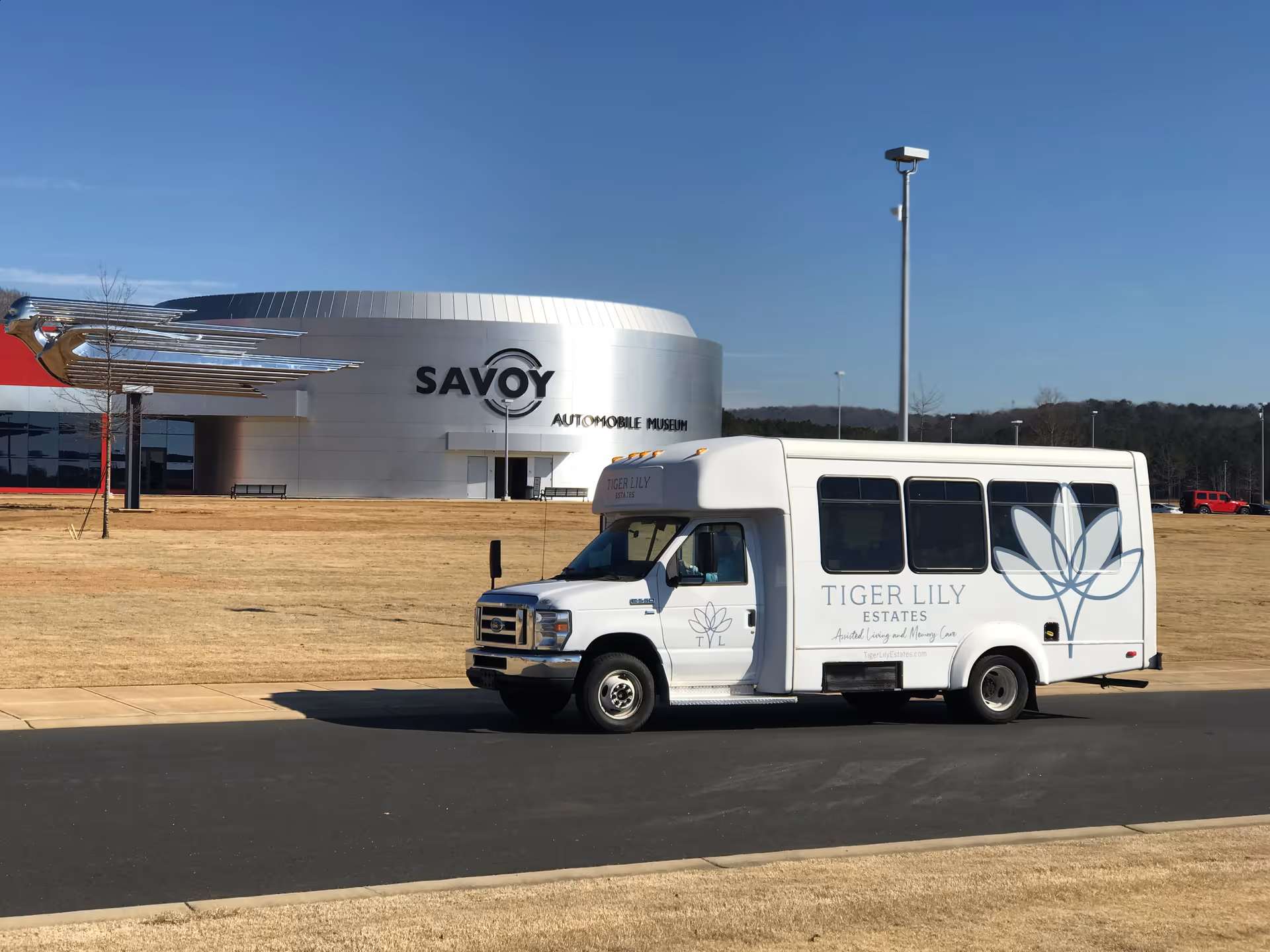 A white shuttle bus with the logo and name 'Tiger Lily Estates Assisted Living and Memory Care' parked on a road in front of the Savoy Automobile Museum building on a clear day.