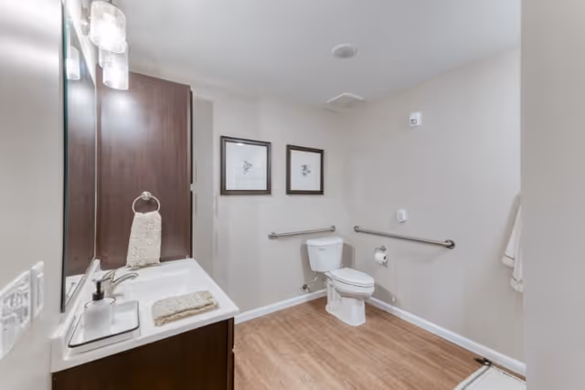 A clean and spacious bathroom with a white toilet equipped with safety grab bars on the walls. There is a dark wood vanity with a white sink, a soap dispenser, and a beige hand towel. Two framed pictures hang on the wall above the toilet. The floor has a wood-like finish and the walls are painted light beige.