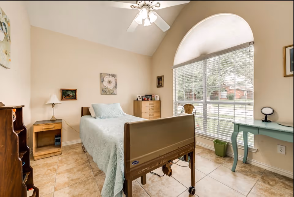 A bright bedroom with a single bed covered in a light blue quilt, a wooden nightstand with a lamp, a chest of drawers, a small desk with a mirror, and a large window with blinds and an arched top letting in natural light.