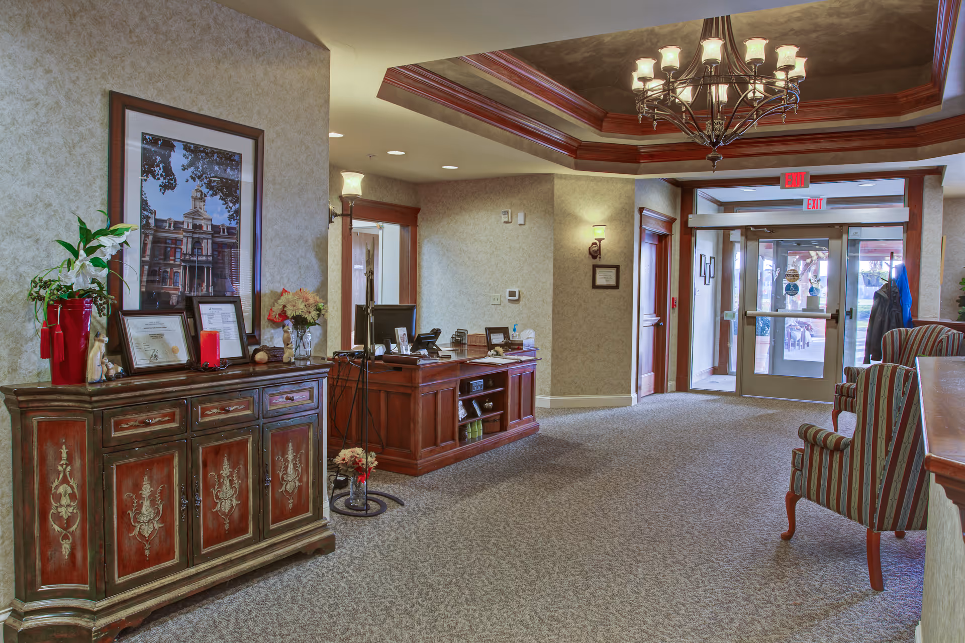 Lobby/reception area with a wooden desk, decorative cabinet, seating, chandelier, and glass entrance doors.