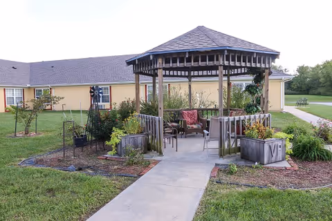 A wooden gazebo with a shingled roof is situated in a garden area with various plants and flowers. There are chairs and a small table inside the gazebo. A concrete pathway leads up to the gazebo, and a yellow building with windows is visible in the background. The surrounding area is grassy with some landscaping and a bench in the distance.