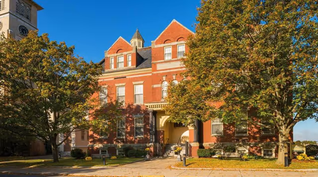 Exterior view of a large, historic brick building with multiple windows and a peaked roof, surrounded by trees with green and yellow leaves under a clear blue sky.