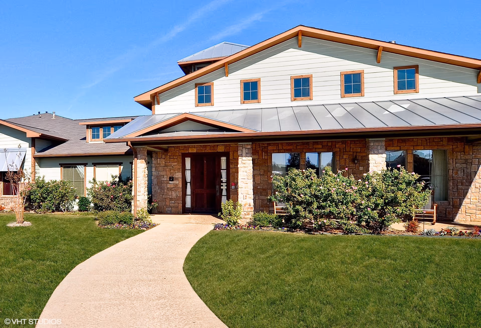 Front exterior view of Pecan Point Assisted Living and Memory Care building with a stone and siding facade, a metal roof, several windows, a walkway leading to the main entrance, and landscaped bushes and grass under a clear blue sky.