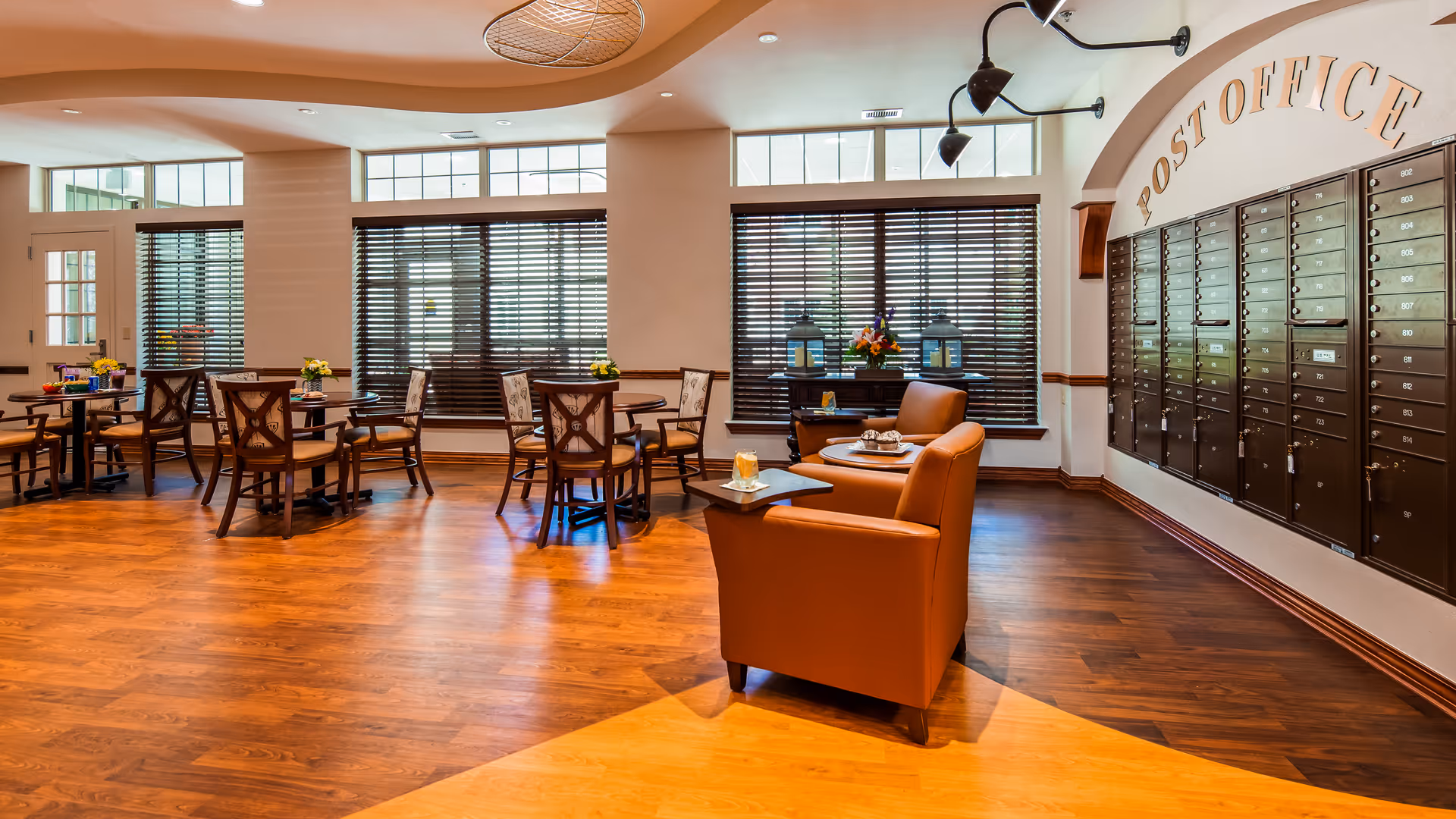 Well-lit senior living common area with tables and chairs, lounge seating, and a wall of mailboxes labeled "Post Office".