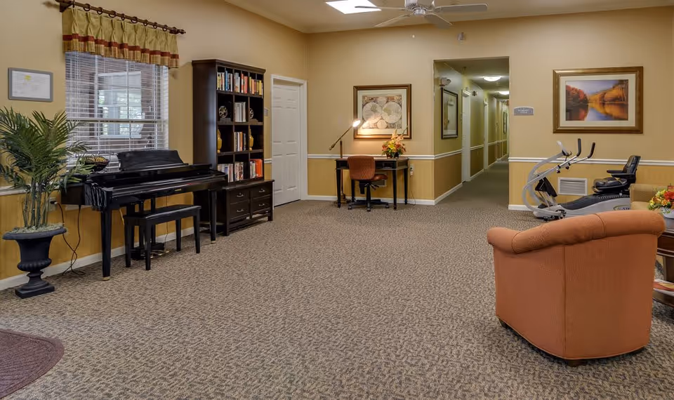 Spacious senior living common room with a piano, bookshelf, desk, exercise equipment, and seating.