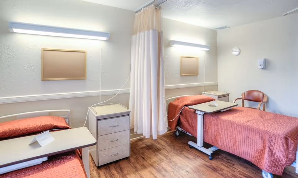 Two medical-style beds with red bedspreads separated by a privacy curtain in a rehabilitation room with overbed tables and wood-look flooring.