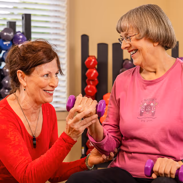 Two elderly women smiling and exercising together indoors, each holding a small purple dumbbell. One woman is assisting the other with her exercise. In the background, there are racks with various colored dumbbells.