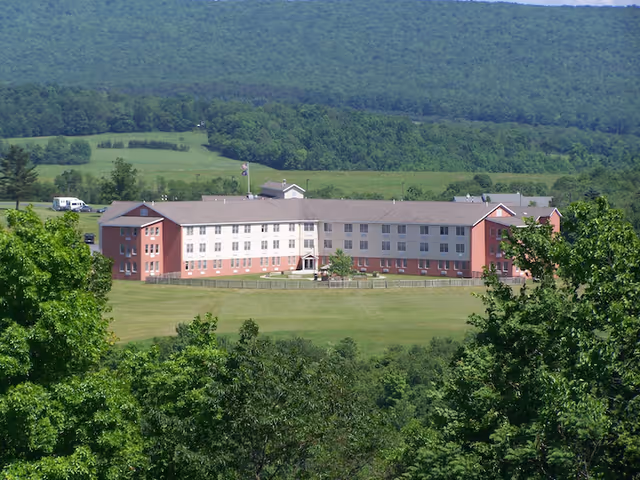 A large, three-story nursing center building with a red and white exterior situated in a spacious green field surrounded by trees and hills in the background.