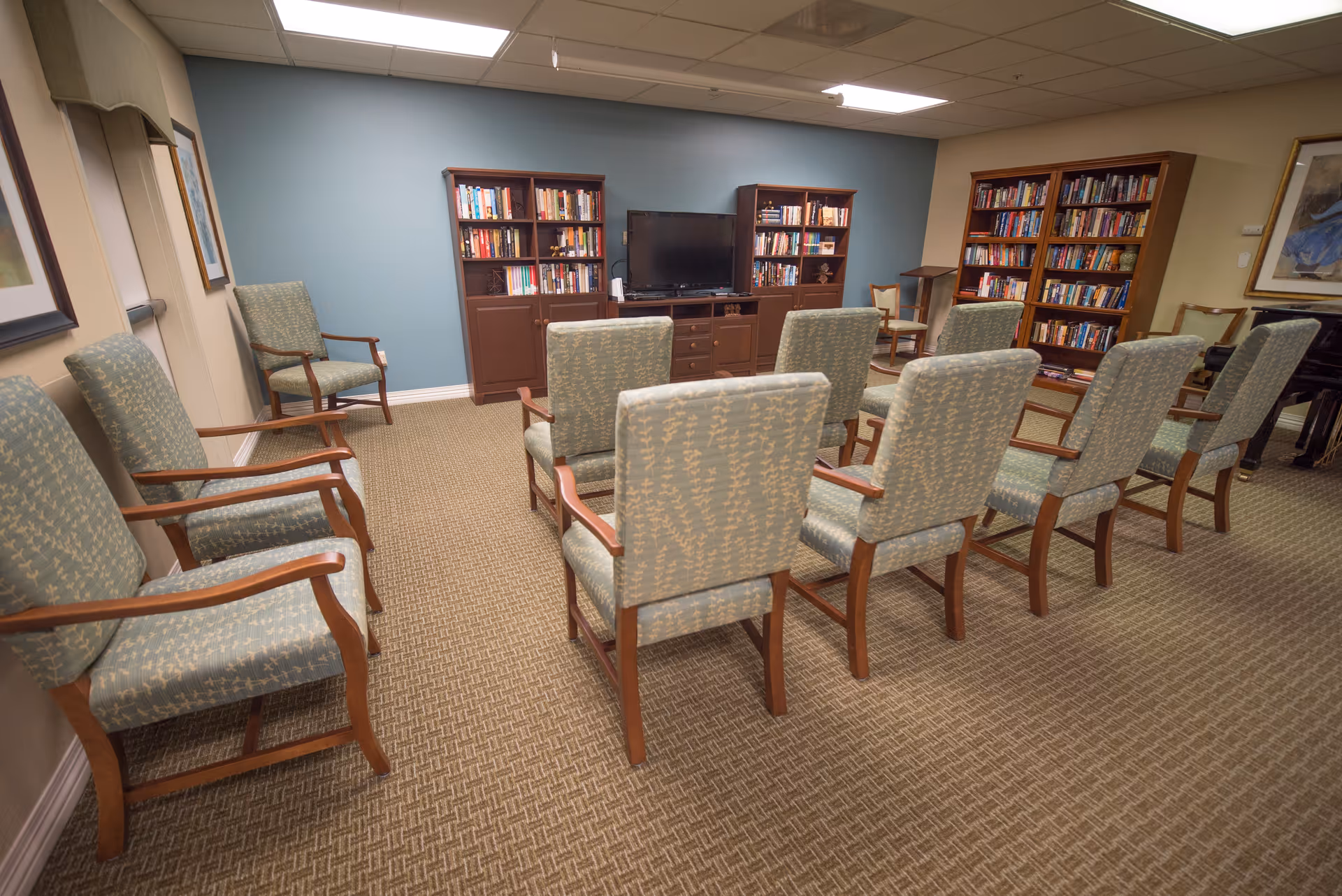 A senior living common room with upholstered chairs arranged facing a TV and bookshelves.