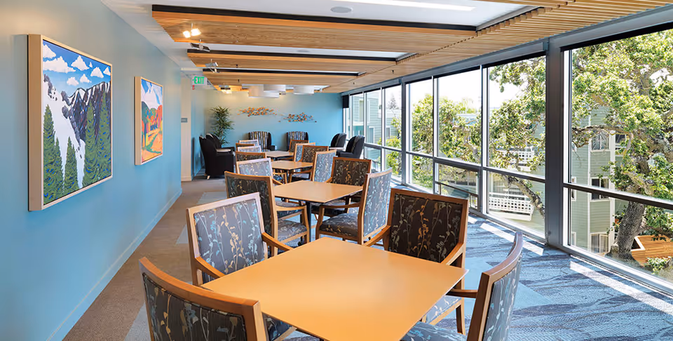 Sunlit dining/lounge area with multiple tables and chairs along floor-to-ceiling windows and artwork on a blue wall.