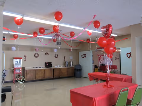A decorated room with red and heart-themed balloons and streamers hanging from the ceiling. Tables covered with red tablecloths and green chairs are arranged in the room. A counter with coffee urns and a popcorn machine is visible along one wall. The room appears to be set up for a celebration or event.