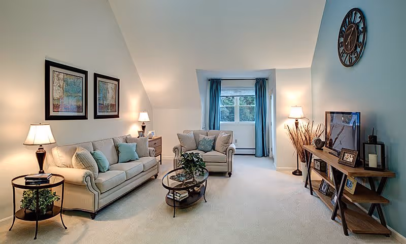 A cozy living room with a beige sofa and armchair adorned with light blue and beige cushions. There are two framed artworks on the wall above the sofa, two table lamps on either side, and a round glass coffee table with a plant on it. A wooden TV stand holds a flat-screen TV, picture frames, and decorative items. A large wall clock is mounted on the teal accent wall, and a window with blue curtains lets in natural light.