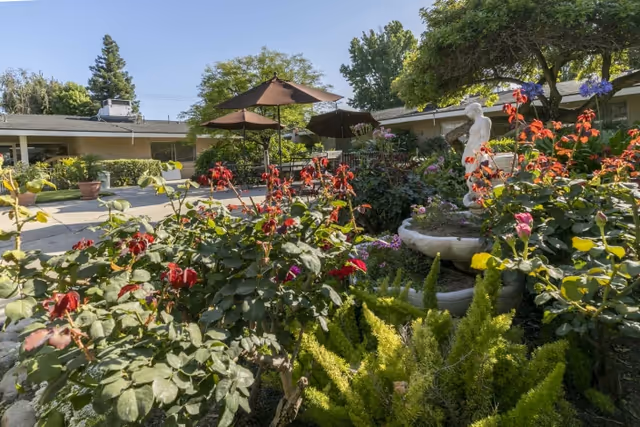 Sunny courtyard garden with flowering plants, a small fountain statue, patio umbrellas, and a surrounding single-story building.