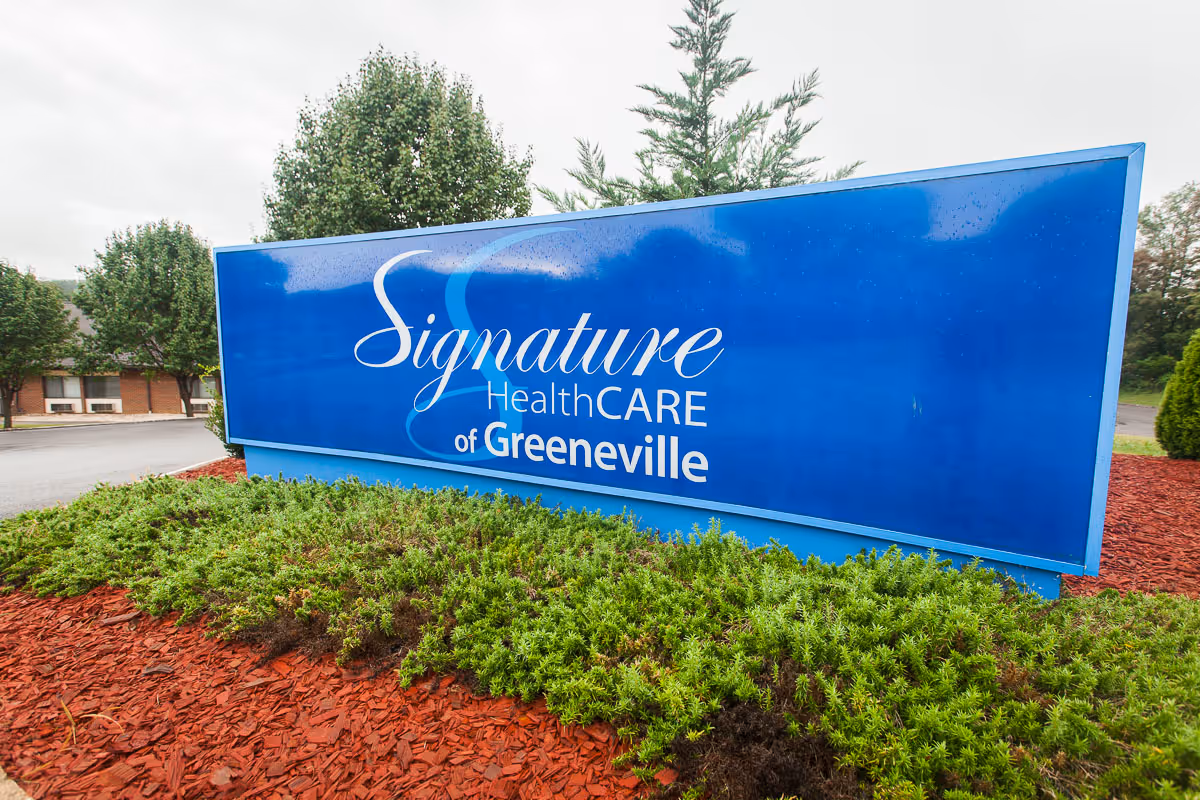 Large blue outdoor sign reading "Signature HealthCARE of Greeneville" set in landscaped mulch and shrubs with trees in the background.