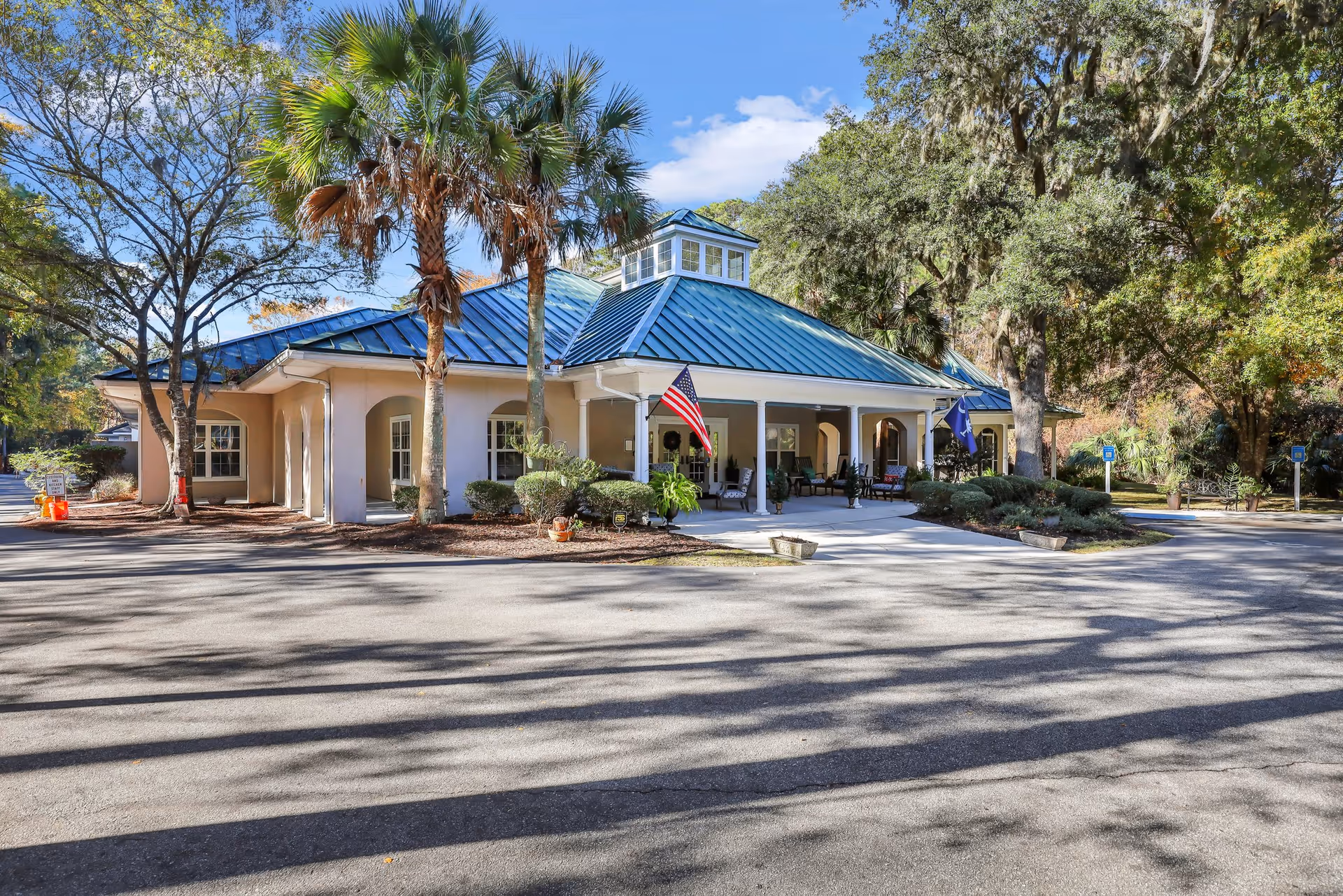 Exterior view of a single-story building with a blue metal roof and a covered porch area. The building is surrounded by trees and landscaping, with an American flag and another flag displayed near the entrance. The sky is clear and blue.