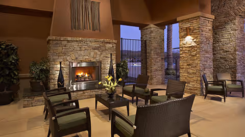 Covered outdoor seating area with stone columns and a lit fireplace surrounded by chairs and a coffee table.