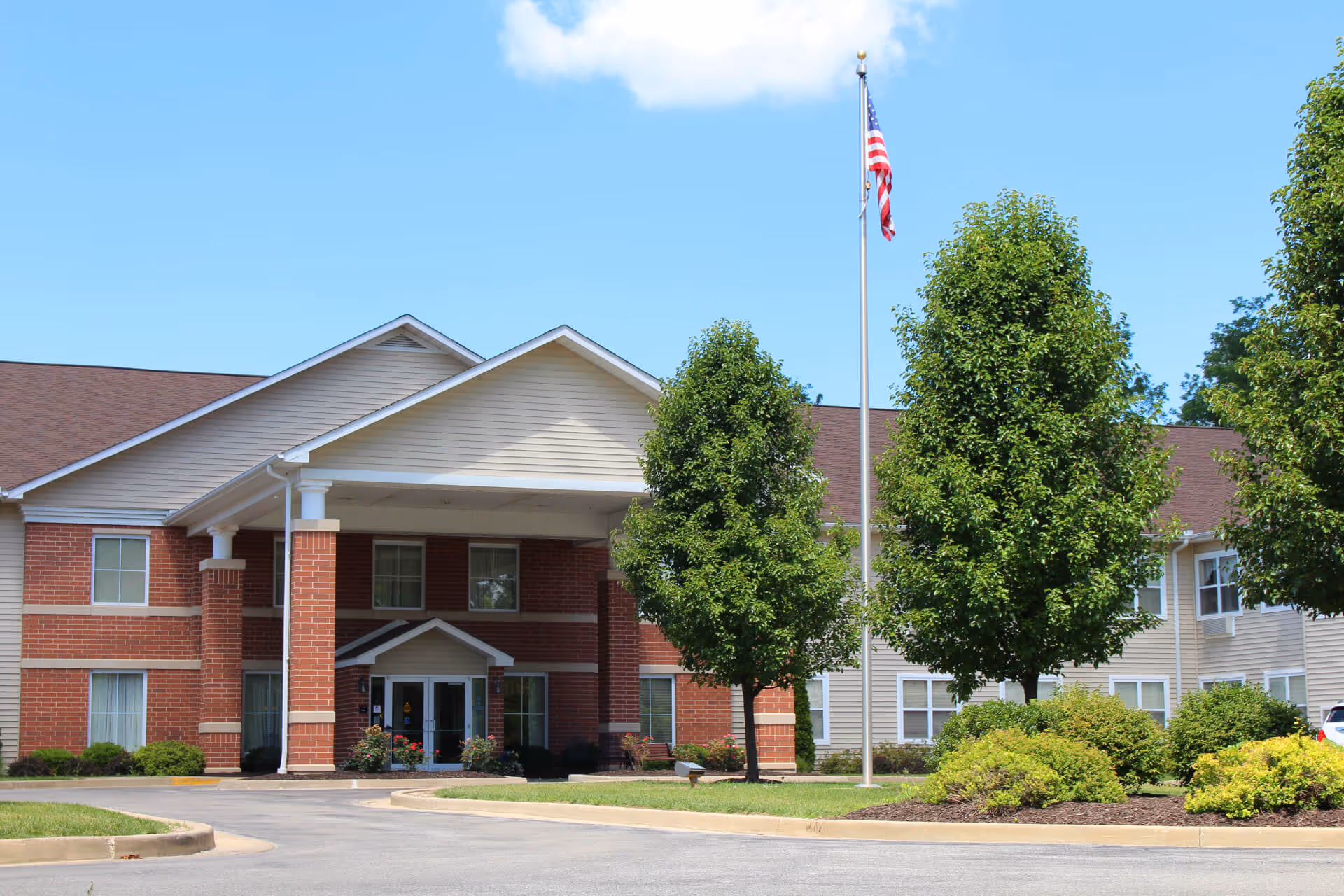 Front entrance of a two-story senior living facility with a covered portico, flagpole, and landscaped grounds.