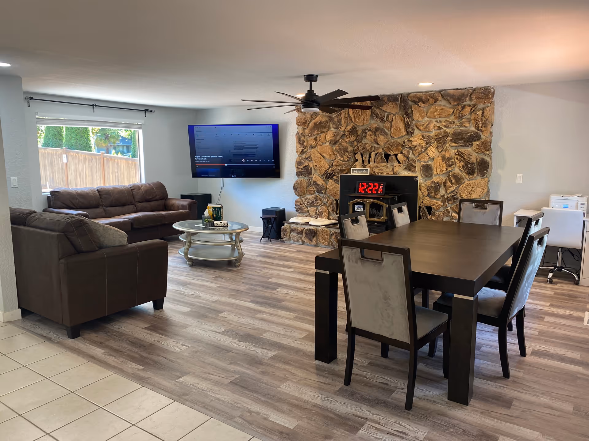 Open living and dining area with brown sofas, a wall-mounted TV, a stone fireplace, and a dark wood dining table on light wood flooring.