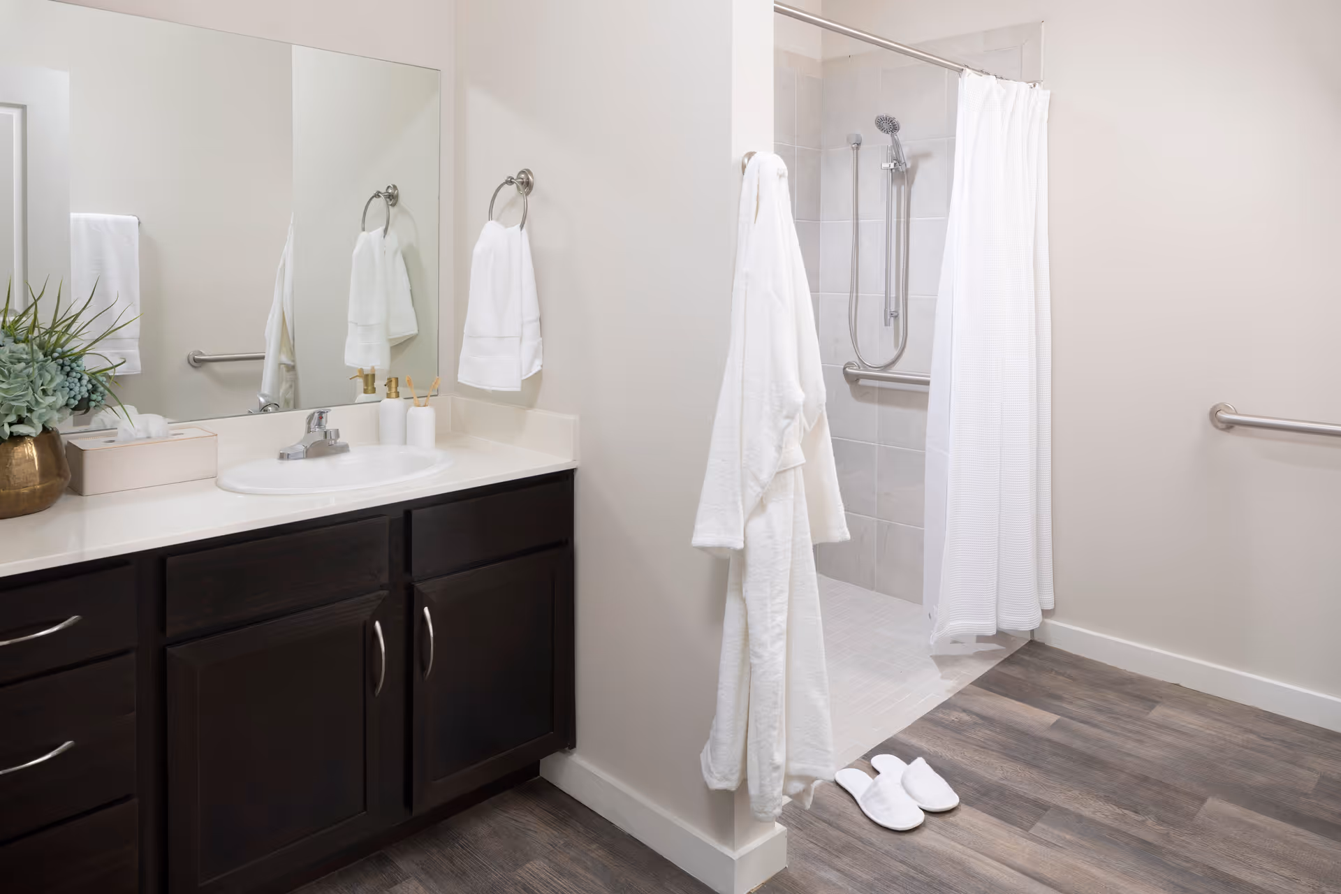 A clean and modern bathroom featuring a dark wood vanity with a white countertop and sink, a large mirror above the sink, a white towel hanging on a ring, a white bathrobe hanging on the wall, white slippers on the floor, and a walk-in shower with a white curtain and grab bars.