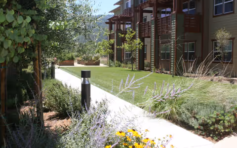 A landscaped outdoor walkway at Merrill Gardens at Gilroy, featuring a concrete path bordered by green grass, flowering plants, shrubs, and trees. On the right side, there is a multi-story building with balconies and windows. The scene is bright and sunny with hills visible in the background.
