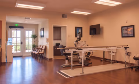 Interior view of a rehabilitation or therapy room in a senior living facility with wooden flooring, parallel bars for walking exercises, an exercise bike, chairs lined up against the wall near a glass exit door, a wall-mounted TV, and framed pictures on the walls.