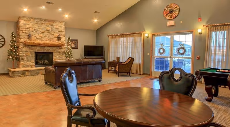 Spacious assisted living common room with a round table and chairs in the foreground, leather sofa facing a stone fireplace, TV, pool table, and double doors.