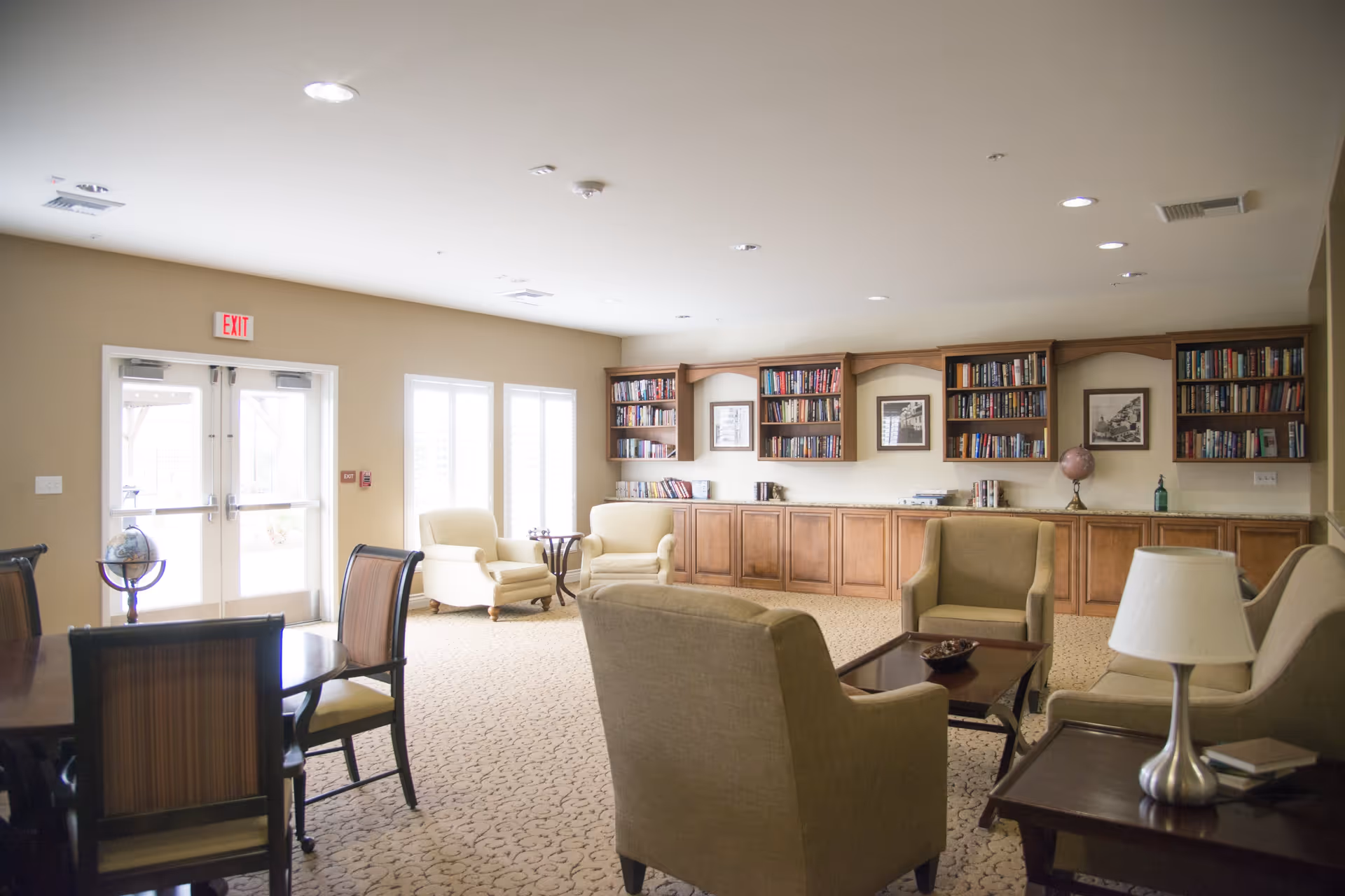 A bright and spacious living room area with beige walls and carpet. The room features multiple comfortable armchairs and a sofa arranged around a wooden coffee table. There are built-in wooden bookshelves filled with books along one wall, with framed pictures hanging above. A table lamp and a globe are also visible, along with a dining table and chairs near a set of double glass doors leading outside.