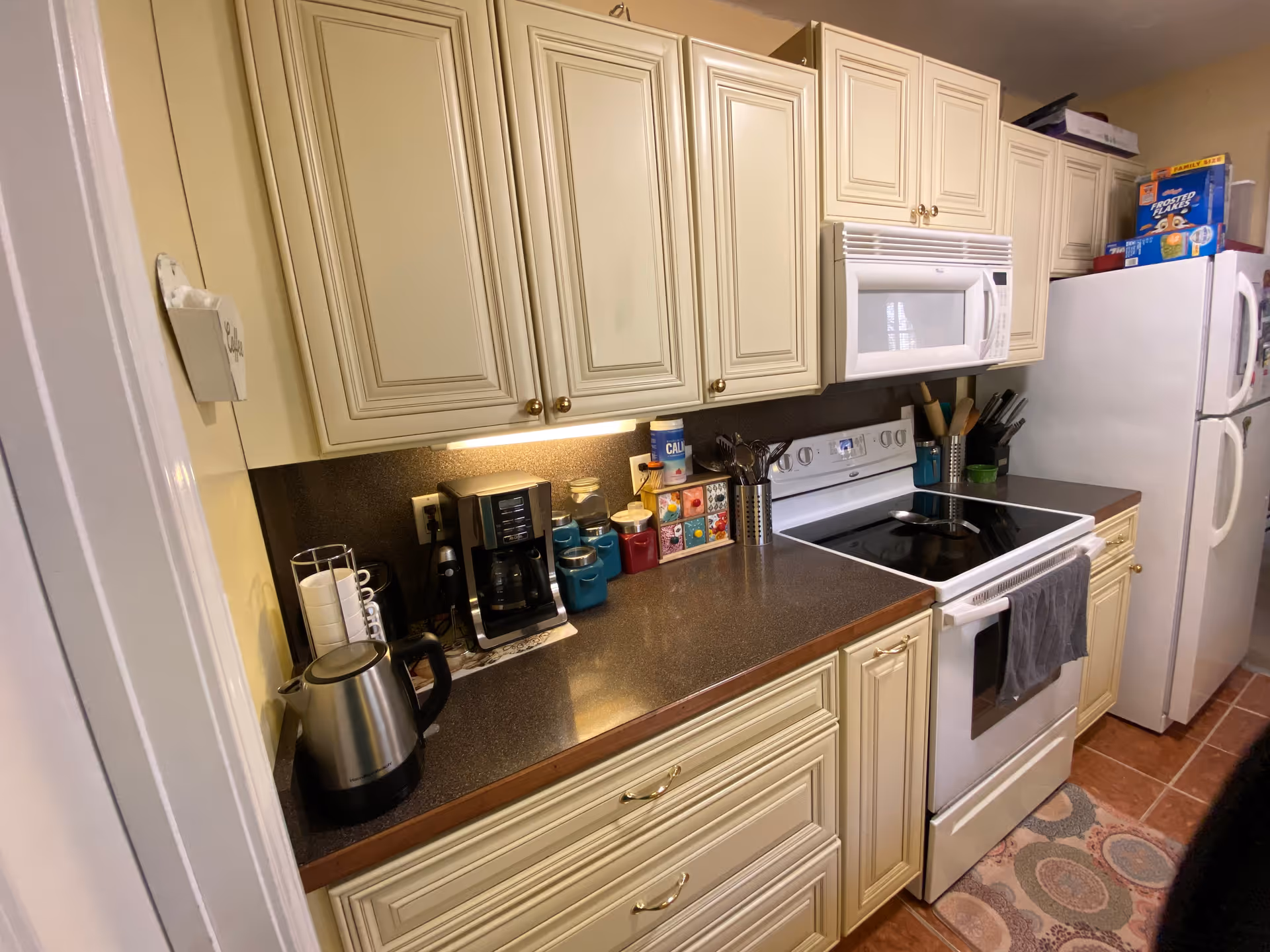 A compact kitchen with cream-colored cabinets, a dark countertop, and various kitchen appliances including a coffee maker, electric kettle, microwave, and stove. There is a white refrigerator with cereal boxes on top and a patterned rug on the tiled floor.