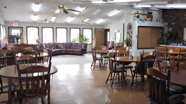 Spacious common room with multiple round wooden dining tables and chairs, ceiling fans, and a lounge area with a purple sectional by windows.