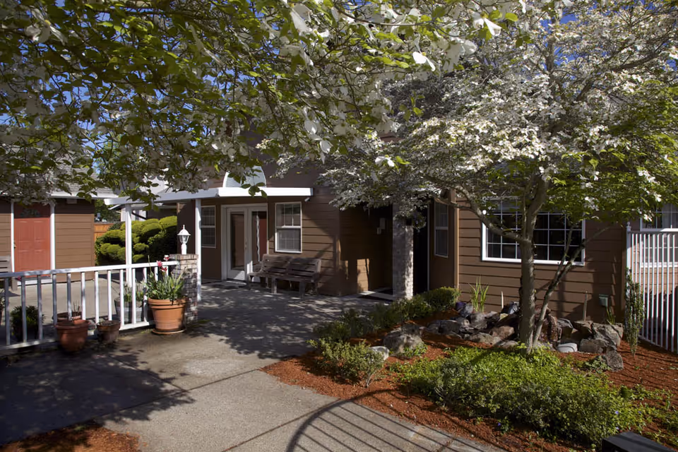 Entrance area of a senior living facility with brown exterior walls, white trim, and a small porch with a bench. The area is shaded by flowering trees and surrounded by well-maintained landscaping including bushes, potted plants, and rocks.
