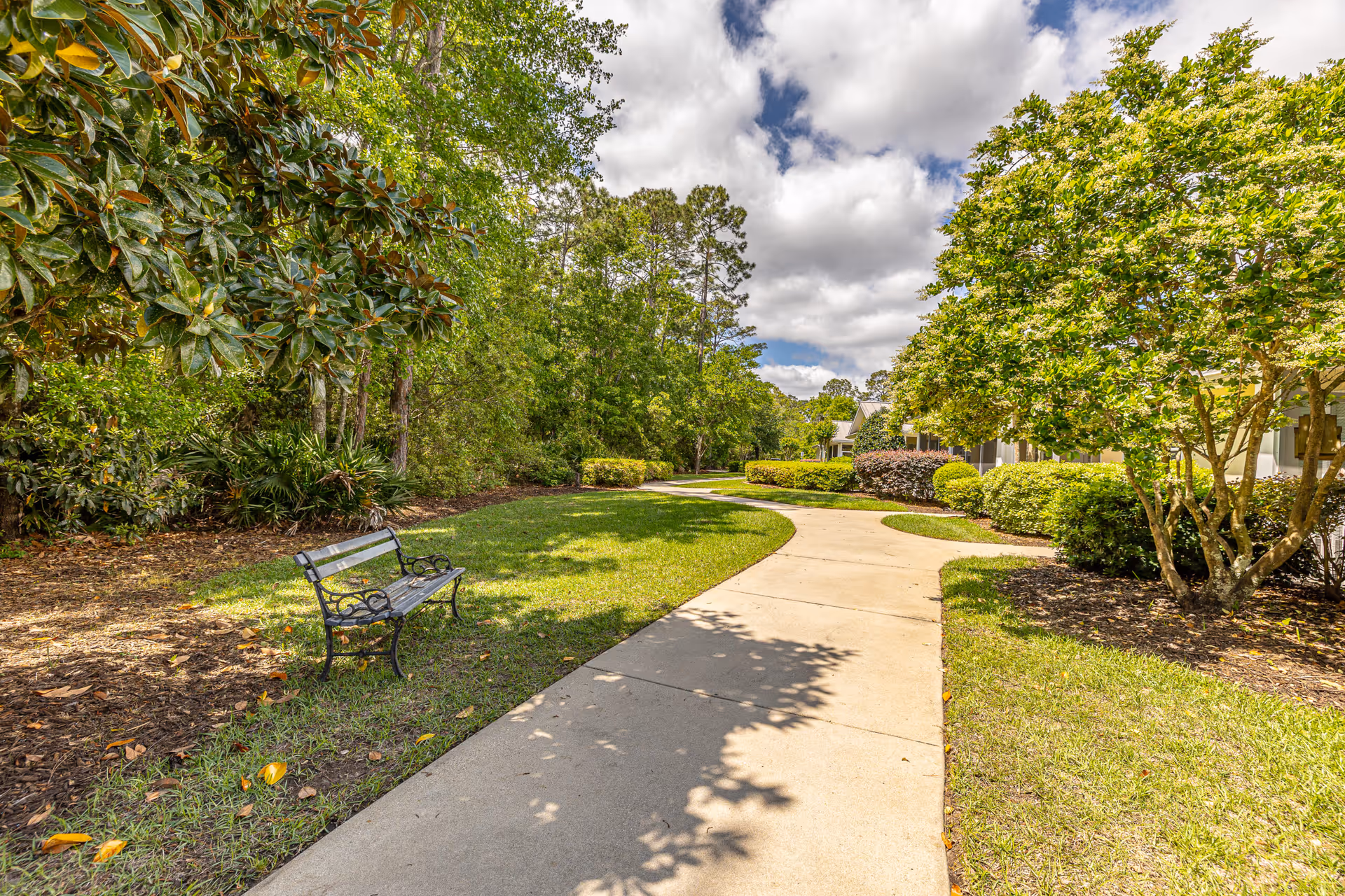 A curved concrete pathway surrounded by well-maintained green grass, bushes, and trees under a partly cloudy sky. A black metal bench is placed on the left side of the path near some trees and mulch. Residential buildings are partially visible on the right side behind the bushes.